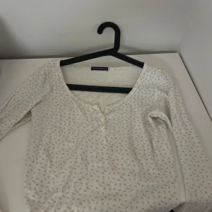 Vit blommig långärmad topp från Brandy Melville - Söt vit långärmad topp från Brandy Melville med små blommor i ljusrosa och grönt. Toppen har knappar framtill och är ribbad i materialet. Perfekt för lager-på-lager och har en feminin, mjuk känsla.