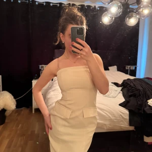 Krämvit bandeauklänning med fickdetaljer - Säljer en stilren krämvit bandeauklänning med raka linjer och dekorativa fickdetaljer framtill. Klänningen har slits bak och är axelbandslös, perfekt för dig som gillar minimalistisk och elegant stil.