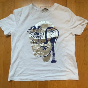 Vit t-shirt med konstnärligt ansikte från Zara - Vit t-shirt från Zara med ett stort konstnärligt tryck av ett ansikte i guld och mörkblått på framsidan. Klassisk rund hals och korta ärmar. Perfekt för dig som gillar unika och kreativa plagg.