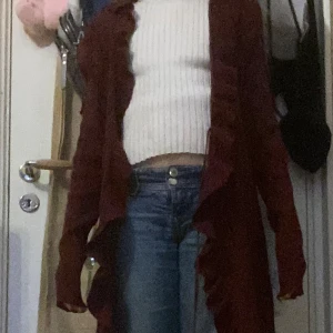 Vinröd kofta med volangkant - Säljer en vinröd, ribbad kofta med långa ärmar och volangkant längs öppningen. Koftan är öppen framtill och har en mjuk, stickad känsla. Perfekt att slänga över en topp för en snygg lager-på-lager-look.