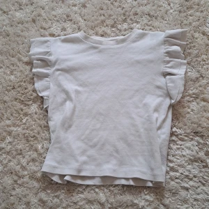 Vit ribbad topp med volangärmar från Gina Tricot - Säljer en söt vit ribbad topp från Gina Tricot i storlek 146/152. Toppen har korta ärmar med volangdetaljer som ger en extra touch. Perfekt till sommaren och enkel att matcha med olika outfits.