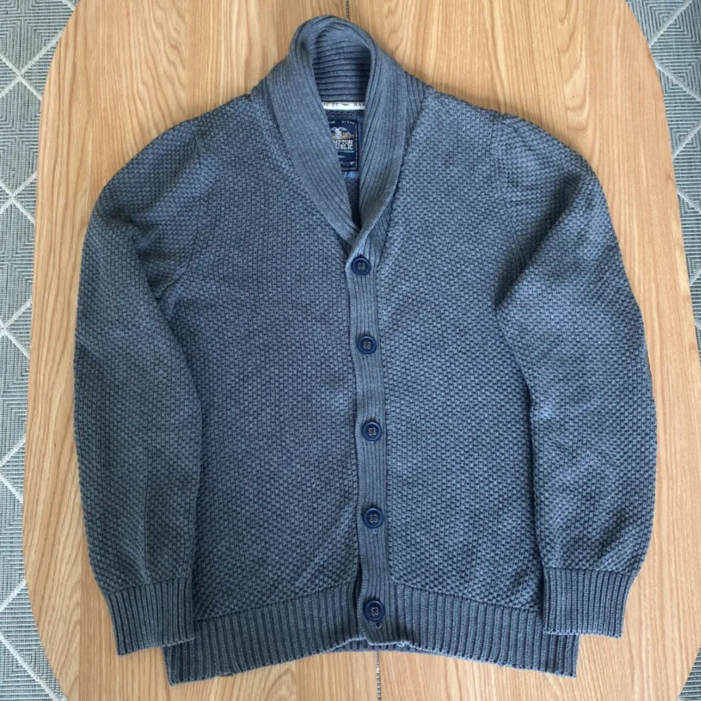 Säljer en grå stickad cardigan med sjalkrage och knäppning framtill. Cardiganen har ribbade muddar och strukturstickat material, perfekt för lager-på-lager. Klassisk och stilren design.. Neuletakit & Villapaidat.