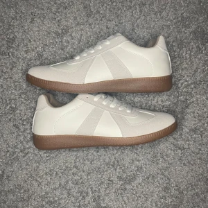 Vita sneakers med beige detaljer och brun sula - Säljer ett par stilrena vita sneakers med beige mockadetaljer och bruna gummisulor. Skorna har snörning och en klassisk, minimalistisk design som passar till många outfits. Perfekta för dig som gillar en clean look.