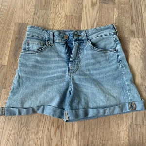 Ljusblå jeansshorts med upprullad kant - Säljer ett par ljusblå jeansshorts med klassisk femficksdesign och upprullad kant nedtill. Shortsen har bälteshällor och knappgylf. Perfekta för varma dagar och enkel att matcha med olika toppar.