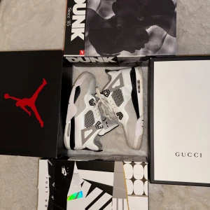 Vita och gråa Air Jordan 4 sneakers - Säljer ett par Air Jordan 4 sneakers i vitt och ljusgrått med svarta detaljer. Skorna har klassisk meshpanel, snörning och den ikoniska Jumpman-loggan på baksidan. Perfekta för dig som gillar streetwear och vill sticka ut med en tidlös modell.