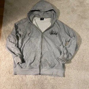 Grå hoodie med dragkedja från Stüssy - Säljer en grå hoodie från Stüssy i storlek S. Tröjan har dragkedja, huva och två fickor framtill. Stüssy-logga finns tryckt både på bröstet och stort på ryggen. Perfekt för en avslappnad streetwear-look.