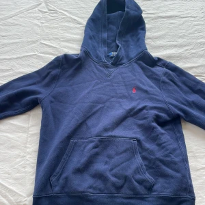 Mörkblå hoodie från Polo Ralph Lauren - Säljer en klassisk mörkblå hoodie från Polo Ralph Lauren med röd broderad logga på bröstet. Tröjan har känguruficka framtill och huva. Perfekt för en avslappnad stil. Storlek passar 158-170