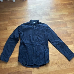 Mörkblå skjorta med knappar - En stilren mörkblå skjorta med klassisk krage och knäppning framtill. Skjortan har långa ärmar och en enkel, tidlös design som passar till många olika tillfällen. Perfekt för dig som gillar en clean och enkel look.