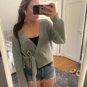 Grå omlott kofta från Chiquelle - Säljer en mysig grå stickad kofta från Chiquelle. Koftan har en v-ringad design och knytband i midjan för en justerbar passform. Perfekt för kyliga dagar och ger en stilren look. Passar till många olika outfits! Är i onesize, men passar ungefär xs-s