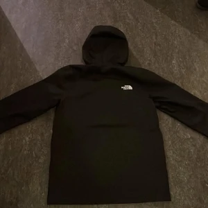 Svart vindjacka från The North Face - Säljer en svart vindjacka från The North Face med huva och dragkedja. Jackan har en stilren design med logga på bröstet och är perfekt för blåsiga dagar. Långärmad modell med praktiska fickor.