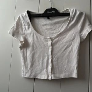 Vit croppad topp med knappar från Divided - Säljer en vit croppad topp från Divided med korta ärmar och knappar framtill. Toppen har en djup rundad halsringning och är tillverkad i mjuk bomull. Perfekt att matcha med jeans eller kjol!