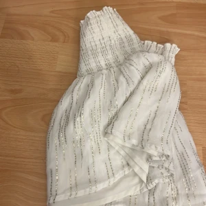 Vit glittrig klänning från Gina Tricot - Supersöt vit klänning från Gina Tricot med glittriga silverränder och smock upptill. Klänningen har en luftig känsla och är perfekt för speciella tillfällen. Materialet är lätt och skimrande, vilket ger en festlig look.