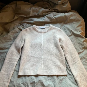 Beige stickad tröja från Zara - Säljer en beige stickad tröja från Zara med rund halsringning och diskret vit söm som detalj framtill och baktill. Tröjan har lång ärm och en rak, något boxig passform. Perfekt för lager-på-lager under kyligare dagar.