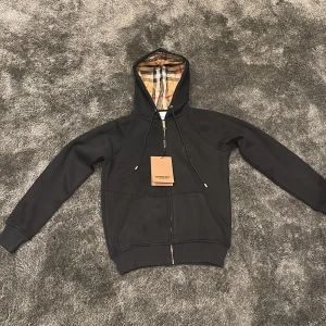 Svart Burberry kofta - Säljer en svart hoodie från Burberry med dragkedja och huva. Insidan av huvan har det ikoniska Burberry-rutiga fodret. Tröjan är i bomull och har ribbade muddar vid ärmslut och nederkant. Perfekt för en stilren och lyxig look. Den är liten i storleken och passar någon med storleken S/XS