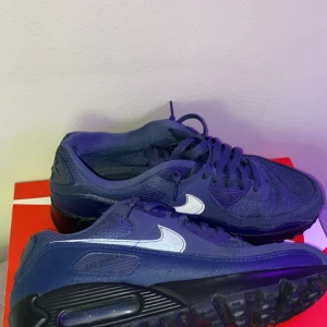 Nike Air Max blå sneakers - Säljer ett par blå Nike Air Max sneakers med vit swoosh och svart sula. Skorna har snörning och en klassisk rund tå. Perfekta för dig som gillar sportig stil och vill ha bekväma skor till vardags.