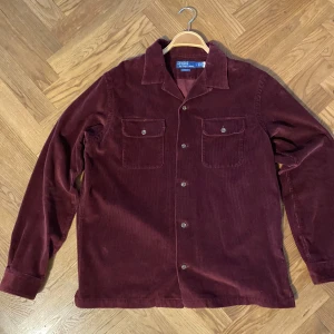 Vinröd manchester overshirt från Polo Ralph Lauren - Säljer en vinröd overshirt i manchester från Polo Ralph Lauren. Skjortan har två bröstfickor med knappar och klassisk krage. Perfekt att bära som lager över en t-shirt. Har endast använts vid 2 tillfällen.