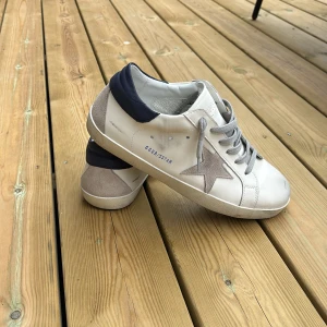 Vita sneakers från Golden Goose med grå stjärna - Snygga vita sneakers från Golden Goose med grå stjärndetalj på sidan och mörkblå häl. Skorna har grå snörning och platt sula, perfekt för en avslappnad stil. Box ingår 