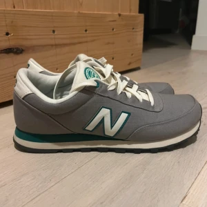 Grå New Balance 501 sneakers - Snygga New Balance 501 sneakers i grått med vita och turkosa detaljer. Skorna har klassisk rund tå, vit sula med turkos rand och vit snörning. Ovandelen är i textil och syntet, perfekt för en clean och sportig look.