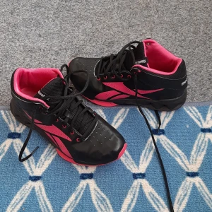 Svarta och rosa Reebok sneakers - Snygga Reebok sneakers i svart med rosa detaljer och innersula. Skorna har snörning, rund tå och en cool sportig design med perforerade partier framtill. Perfekta för dig som gillar att sticka ut med färg och stil på gymmet eller i vardagen.