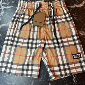 Rutig shorts från Burberry - Snygga shorts från Burberry i klassiskt beige, svart, vit och röd rutig design. De har elastisk midja med snörning och en patch med Burberry-logga framtill. Perfekta för sommaren och har en modern passform.