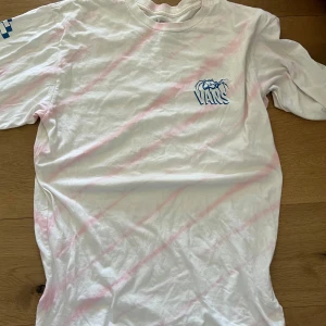 Vit och rosa tröja från Vans - Säljer en vit tröja från Vans med ljusrosa diagonala ränder och blå tryckdetaljer fram och bak. Tröjqn har rund hals och korta ärmar. Perfekt för dig som gillar streetwear och snygga prints.