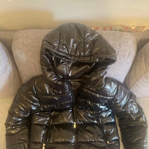Svart glansig pufferjacka med huva - Säljer en svart, glansig pufferjacka med stor huva och dragkedjor på fickorna. Jackan har en quiltad look och är perfekt för kalla dagar. Den har långa ärmar och en riktigt snygg streetstil som sticker ut.