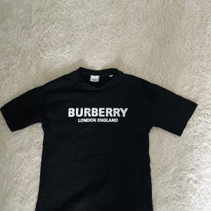 Svart t-shirt från Burberry - Svart t-shirt från Burberry med vit text 'BURBERRY LONDON ENGLAND' tryckt på bröstet. Klassisk rund halsringning och korta ärmar. Perfekt för en stilren och enkel look.