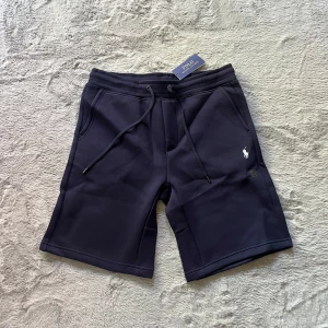 Mörkblå shorts från Polo Ralph Lauren - Snygga mörkblå shorts från Polo Ralph Lauren med vit broderad logga på benet. Modellen har snörning i midjan och två sidofickor. Tillverkade i mjukt material och har en slim fit passform.