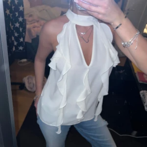 Vit volangtopp från Zara - Säljer en vit ärmlös topp från Zara med hög hals och volangdetaljer längs framsidan. Toppen har en snygg v-ringning och knäpps i nacken. Perfekt att styla med jeans eller kjol. Aldrig använd