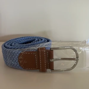 Ljusblått flätat bälte - Säljer ett ljusblått flätat bälte med bruna skinn-detaljer och silverspänne. Bältet har en modern look och passar perfekt till jeans eller chinos. Snygg kontrast mellan det blå bandet och de bruna detaljerna. Är elastiskt! 