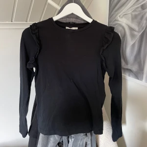 Svart långärmad topp med volang från MANGO - Svart långärmad topp från MANGO med volangdetaljer längs axlarna. Enkel och stilren design med rund halsringning. Perfekt att matcha med jeans eller kjol för en snygg look.