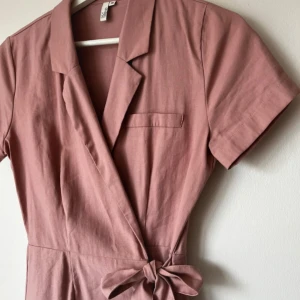 Rosa omlottklänning med knytband - Säljer en rosa kortärmad omlottklänning med krage och knytband i midjan. Klänningen har en bröstficka som detalj och är tillverkad i ett mjukt bomullsliknande material. Perfekt för en avslappnad och stilren look.