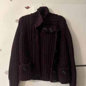 Mörklila stickad kofta från BOSS - Säljer en chunky mörklila stickad kofta från BOSS med stora svarta knappar framtill och på fickorna. Koftan har hög krage och grovstickat mönster, perfekt för lager-på-lager. Snygg och mysig till kalla dagar.