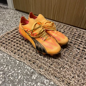 Puma fotbollsskor i orange och gul - Säljer ett par snygga fotbollsskor från Puma i orange och gul med svarta detaljer. Skorna har snörning och dobbar för spel på gräs. Perfekta för dig som vill sticka ut på planen!