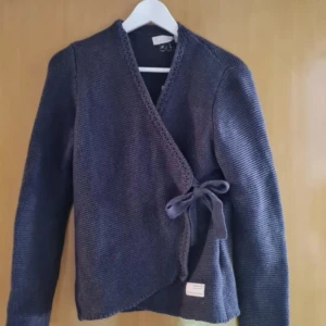 Mörkblå omlottkofta med rosett - Säljer en mörkblå stickad omlottkofta med knytband i sidan. Mycket bra skick och väldigt fin! Skriv vid frågor eller pris förslag!