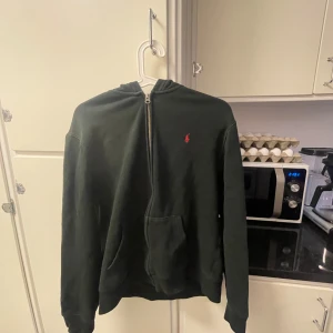 Svart hoodie från Polo Ralph Lauren - Klassisk svart hoodie från Polo Ralph Lauren med dragkedja och röd broderad logga på bröstet. Tröjan har huva och framficka, perfekt för en avslappnad stil.