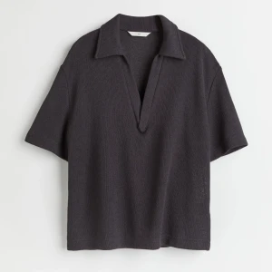 Stickad t shirt - Säljer en mörkgrå stickad tröja från H&M med korta ärmar och djup v-ringning. Tröjan har en klassisk krage och är tillverkad i ett mjukt, ribbat material. Perfekt för en avslappnad men stilren look.