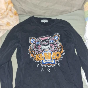Svart Kenzo tröja med tigerbroderi - Säljer en svart långärmad tröja från Kenzo med det ikoniska tigerbroderiet och texten 'KENZO PARIS' framtill. Tröjan har rund hals och är i mjukt material, perfekt för en cool och trendig look.