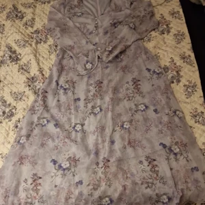Ljuslila blommig långklänning - Helt ny ej använd.Säljer en romantisk långklänning i ljuslila nyans med blommigt mönster i lila, rosa och vitt. Klänningen har långa ärmar, v-ringning och knappar framtill. Perfekt för sommarens tillfällen och midsommarfirande.