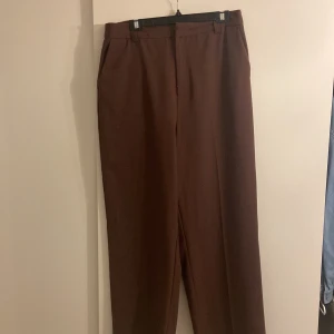 Bruna kostymbyxor från Gina Tricot - Säljer ett par bruna kostymbyxor från Gina Tricot i storlek XL. Byxorna har raka ben, hällor för bälte och diskreta fickor bak. Perfekta för en stilren och klassisk look.