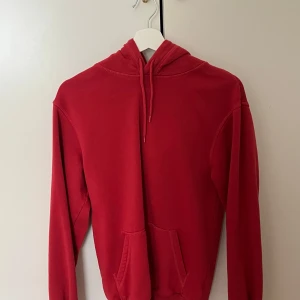 Röd hoodie i bomull - Säljer en enkel röd hoodie med huva och känguruficka framtill. Tröjan har långa ärmar och ribbade muddar vid ärmslut och nederkant. Perfekt för en avslappnad stil.