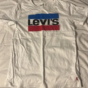Vit t-shirt från Levi's med logga - Säljer en klassisk vit t-shirt från Levi's med stor färgglad logga i blått, svart och rött på bröstet. T-shirten har rund hals och korta ärmar. Perfekt nu inför sommaren.