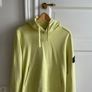 Ljusgul hoodie från Stone Island - Säljer en ljusgul hoodie från Stone Island med klassisk patch på ärmen. Tröjan har huva med snörning och en stor magficka. Tillverkad i bomull och har en clean, stilren look som passar perfekt till streetwear.