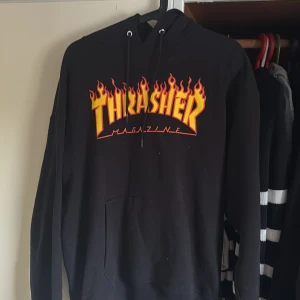 Svart Thrasher hoodie med flames - Svart hoodie från Thrasher med ikoniskt flames-tryck i gult och rött på bröstet. Klassisk huva med snörning och känguruficka framtill. Perfekt streetwear-vibe och riktigt skön passform. Materialet är mjuk bomullsmix.