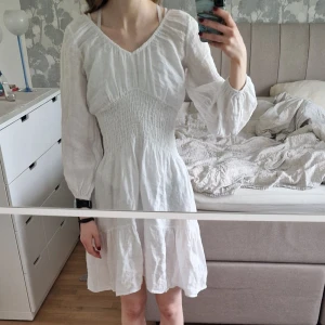 Vit kort klänning i 100%  linne - Superfin vit klänning i linne med v-ringning, smockad midja och långa puffärmar. Den har en flowy kjol med volang och är perfekt för varma dagar. Perfekt för student, skolavslutning eller när som helst en fin sommardag. Köpt på Chelsea i Göteborg.