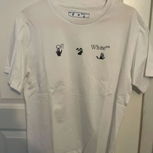 Vit Off-White t-shirt med blått tryck - Säljer en vit t-shirt från Off-White med svart logga och handgrafik framtill. Baksidan har ett stort blått och svart grafiskt tryck i Off-Whites klassiska korsdesign. Skön passform och tillverkad i mjuk bomull. Perfekt för dig som gillar streetwear vibes.