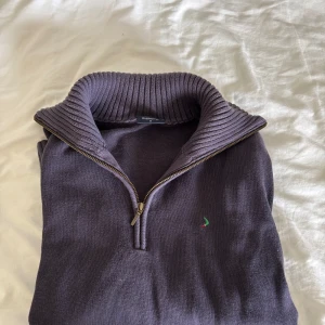 Mörklila stickad half zip-tröja Boomerang - Snygg mörklila stickad tröja från Boomerang med ribbad hög krage och half zip-dragkedja. Liten grön logga broderad på bröstet. Perfekt för lager-på-lager och chillare dagar. Långärmad och med breda ribbade muddar vid ärmslut och nederkant.