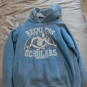 Blå hoodie från Reckless Scholars - Säljer en blå hoodie från Reckless Scholars med tryck på bröstet och stor ficka framtill. Tröjan har huva med dragsko och är tillverkad i mjukt material, perfekt för en avslappnad stil.