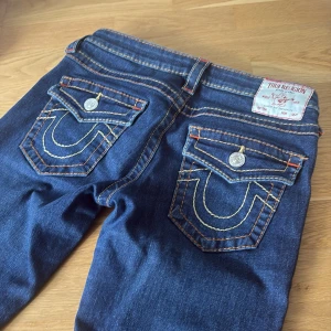 Mörkblå True Religion jeans med kontrastsömmar - Säljer ett par mörkblå jeans från True Religion med ikoniska bakfickor och kontrastsömmar i orange och gul. Byxorna har bootcut-modell och klassisk femficksdesign. Perfekta för dig som gillar detaljer och snygg passform. Säljer då de inte passar❤️