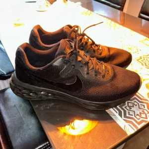 Svarta Nike sneakers - Säljer ett par helsvarta Nike sneakers med mesh-ovandel och platt sula. Skorna har snörning och en diskret Nike-logga på sidan. Perfekta för vardagsbruk eller träning.